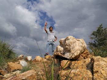 Low angle view of man gesturing while standing on rocks against cloudy sky