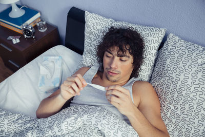 High angle view of young man holding thermometer on bed at home