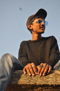 Low angle view of boy wearing sunglasses sitting on retaining wall against sky
