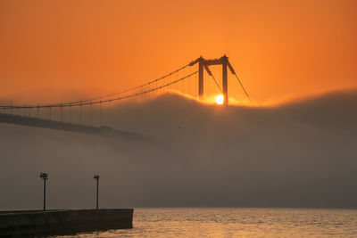 Silhouette bridge over sea against orange sky