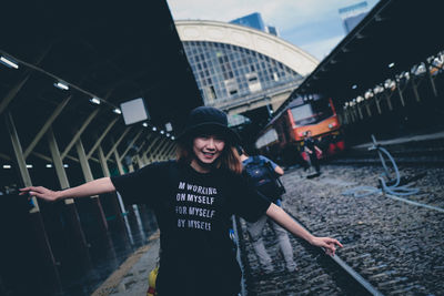 Portrait of smiling young woman with arms outstretched standing on railroad station platform