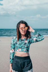 Young woman wearing sunglasses while standing at beach against sky during sunny day