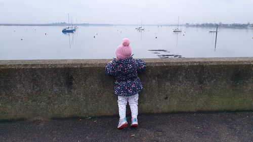 Rear view of girl in warm clothes standing against wall at sea