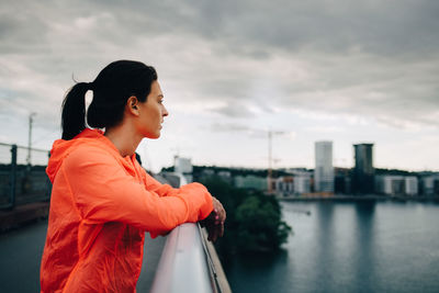 Side view of female athlete in raincoat looking away while standing on footbridge over sea