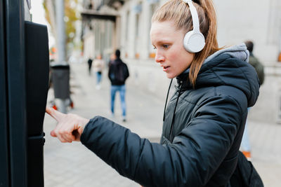 Side view of young woman using parking meter while standing on footpath