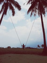 Rear view of boy swinging by palm trees against sky