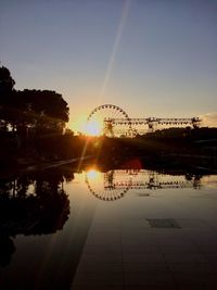 Silhouette ferris wheel by river against sky at sunset