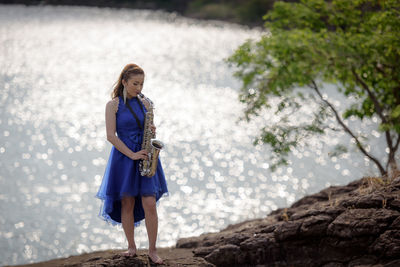 Young bride playing saxophone on rock formation by lake