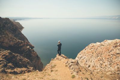 Rear view of man standing on cliff by sea against clear sky