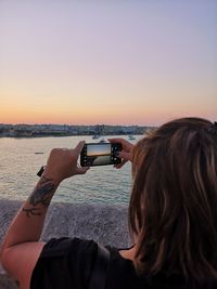 Midsection of woman photographing sea against sky during sunset