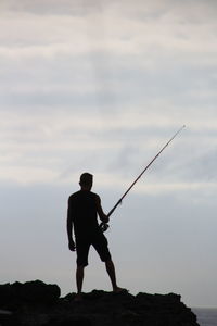 Silhouette man fishing on rock against sky