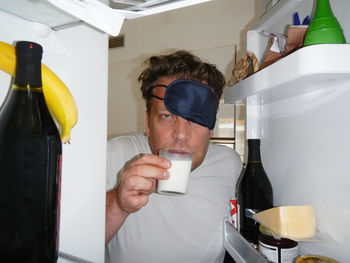 Portrait of tired man wearing sleep mask drinking milk from refrigerator