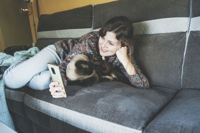 Smiling woman using smart phone while lying on cat at home