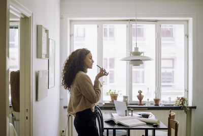 Side view of female architect talking on speaker phone while standing near table at home