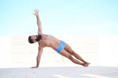 Full length of shirtless man practicing yoga against sky