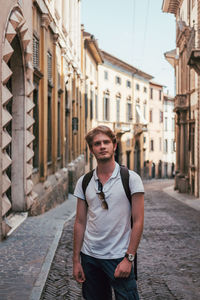 Portrait of young man standing at alley amidst buildings in city