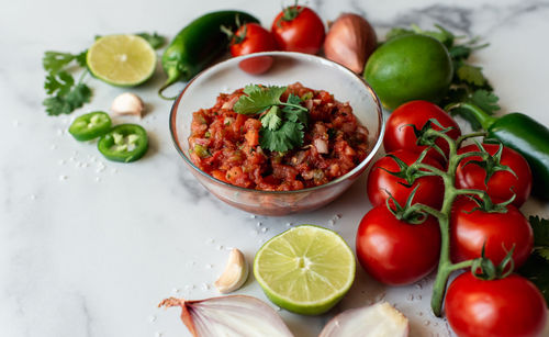 Bowl of fresh salsa with the ingredients on white marble counter.