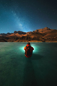 A young man with a backpack sits on the ice of a frozen lake at night in winter and looks 