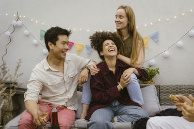 Cheerful male and female friends laughing together while sitting on sofa at party in decorated balcony