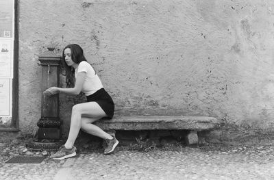 Full length of woman drinking water while sitting on seat against wall