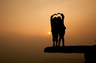 Silhouette of two women making heart shape with hands while standing on rock against sky during sunset