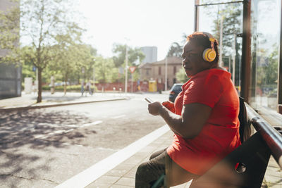 Woman holding smart phone listening music through wireless headphones sitting on bench