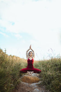 Young woman in red sporty wear standing on toes in lotus pose with hands clasped above head among green tall grass