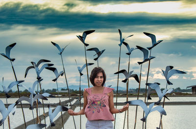 Portrait of mid adult woman standing by sculptures by lake against cloudy sky during sunset