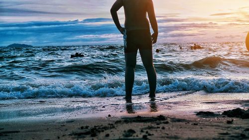 Low section of man standing on shore at beach against sky during sunset