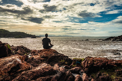 Rear view of man sitting on rock by sea against sky