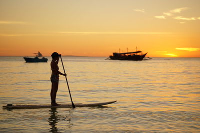 Silhouette woman paddleboarding in sea against orange sky