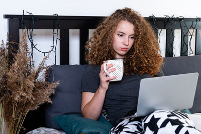 Young woman holding coffee cup while using laptop at home