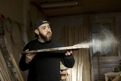 Carpenter blowing sawdust from plank at workshop