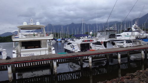 Boats moored by pier in harbor at lake against cloudy sky