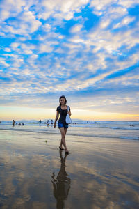 Young woman walking at beach against cloudy sky during sunset