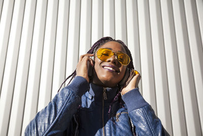 Happy young african american female with afro braids dressed in blue jacket and stylish sunglasses enjoying music through earphones while chilling in sunlight against striped wall