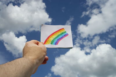 Low angle view of hand holding multi colored rainbow on paper against cloudy sky