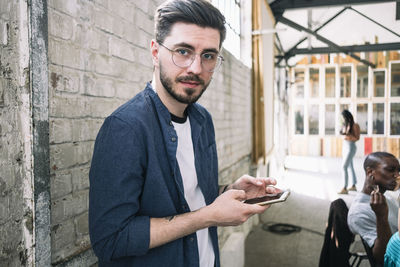 Portrait of confident young businessman standing with smart phone against brick wall at creative workplace