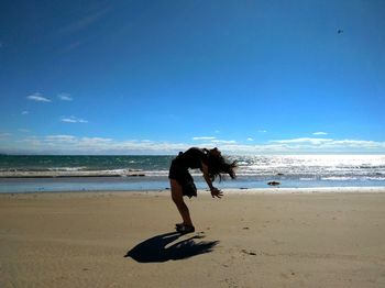 Playful young woman bending over backwards at beach against blue sky
