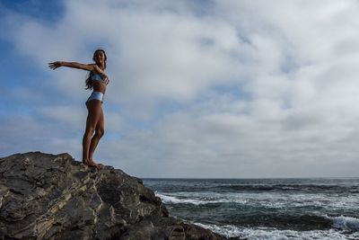 Low angle view of woman with arms outstretched standing on rock by sea against cloudy sky