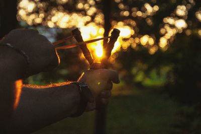 Cropped image of man aiming with slingshot against trees during sunset