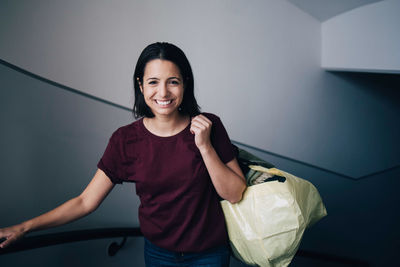 Portrait of smiling woman carrying bag while standing by wall