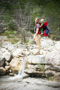 Young man hiking with his baby sitting in the backpack. family is watching small waterfall in forest