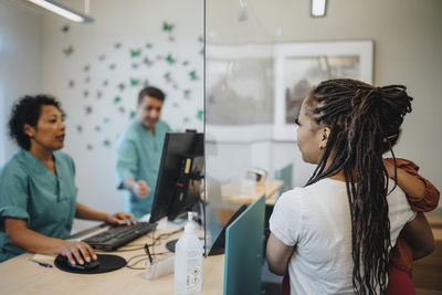 Mother carrying daughter talking with receptionist through glass partition at reception desk in hospital