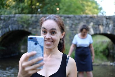 Close-up of cheerful woman taking selfie against bridge over river