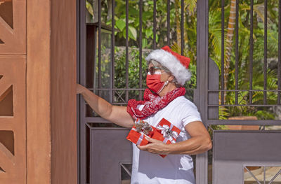 Senior woman wearing mask holding christmas gift standing by door