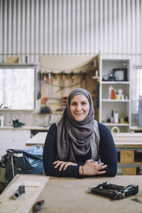 Portrait of smiling female carpenter wearing headscarf leaning on workbench