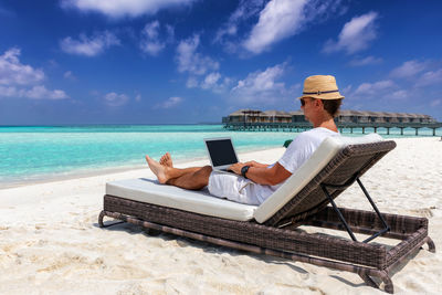 Man using laptop while sitting on lounge chair at beach against sky