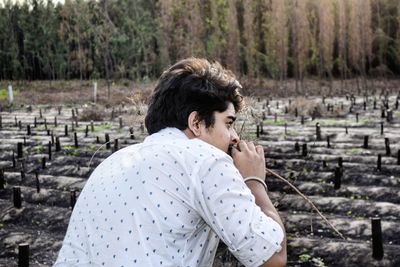 Side view of young man looking away while crouching on field against trees