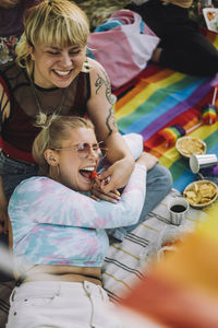 Happy non-binary person feeding strawberry to female friend lying on picnic blanket